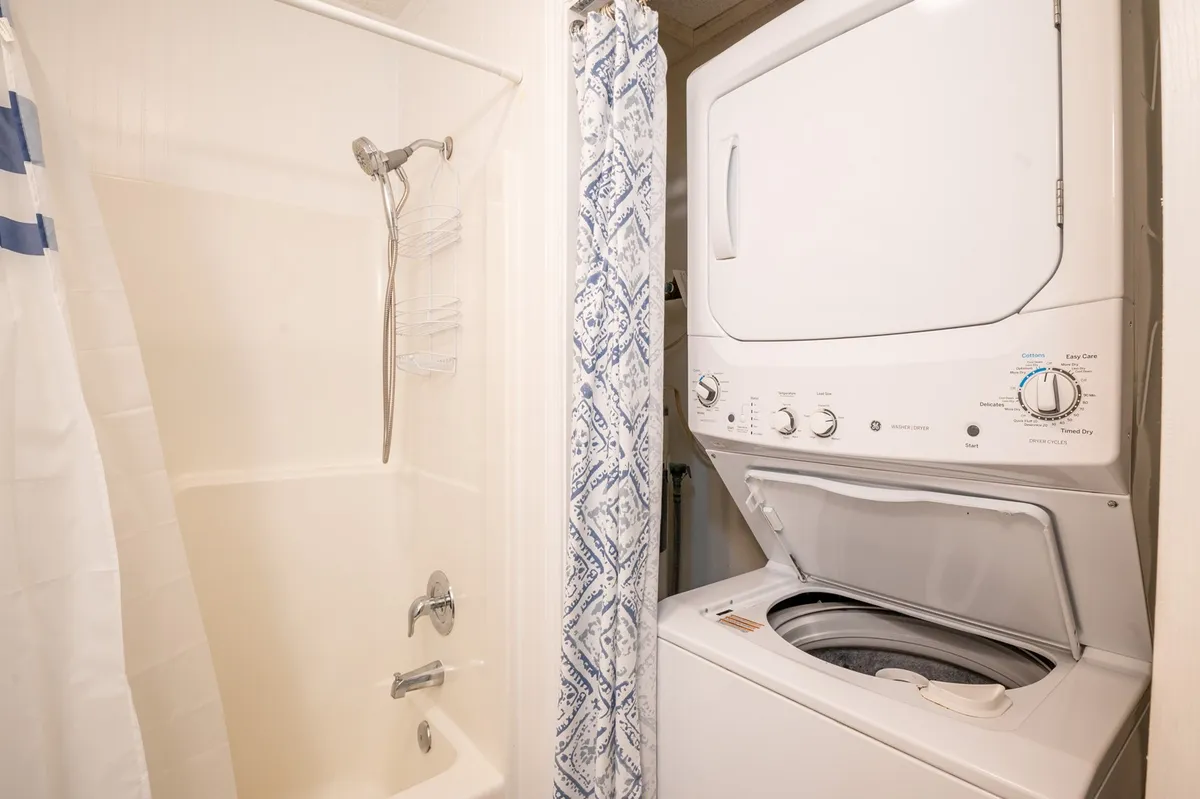 A practical and inviting bathroom space featuring a washing machine tucked beside a shower with a decorative curtain. The setup is clean and accessible, embodying the convenience of in-unit laundry and a spacious hallway bath for guests.