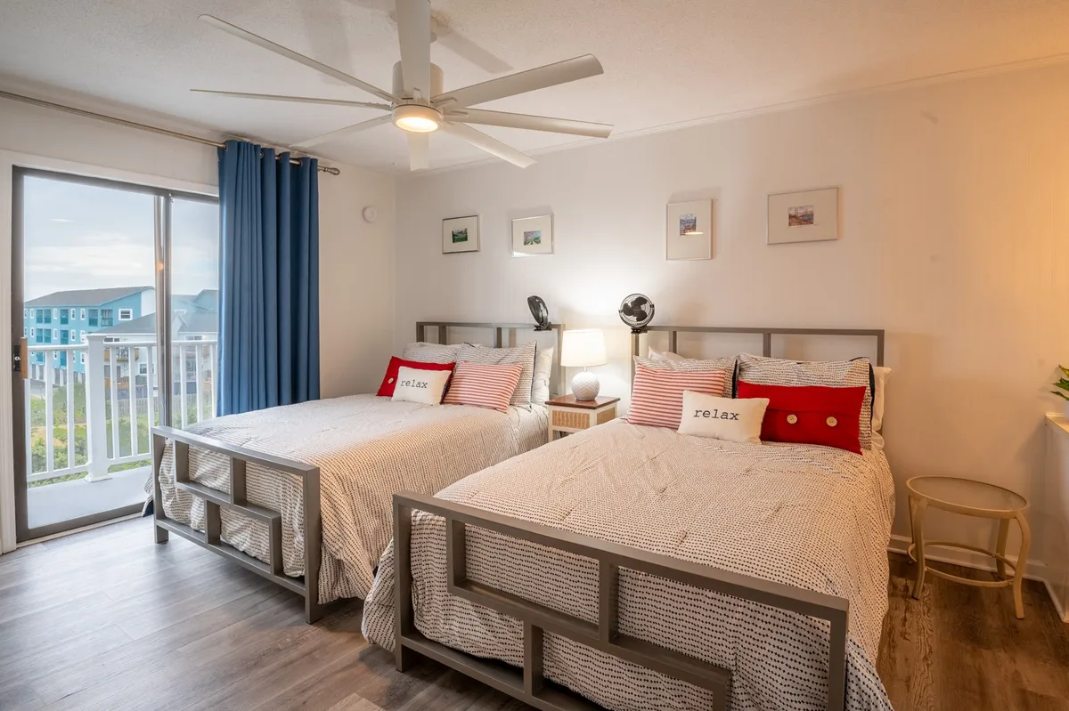 A serene bedroom featuring two beds with decorative pillows, a large window with a view, a ceiling fan, and a nightstand with a lamp. The window draws in natural light and suggests the possibility of relaxing with ocean breezes.