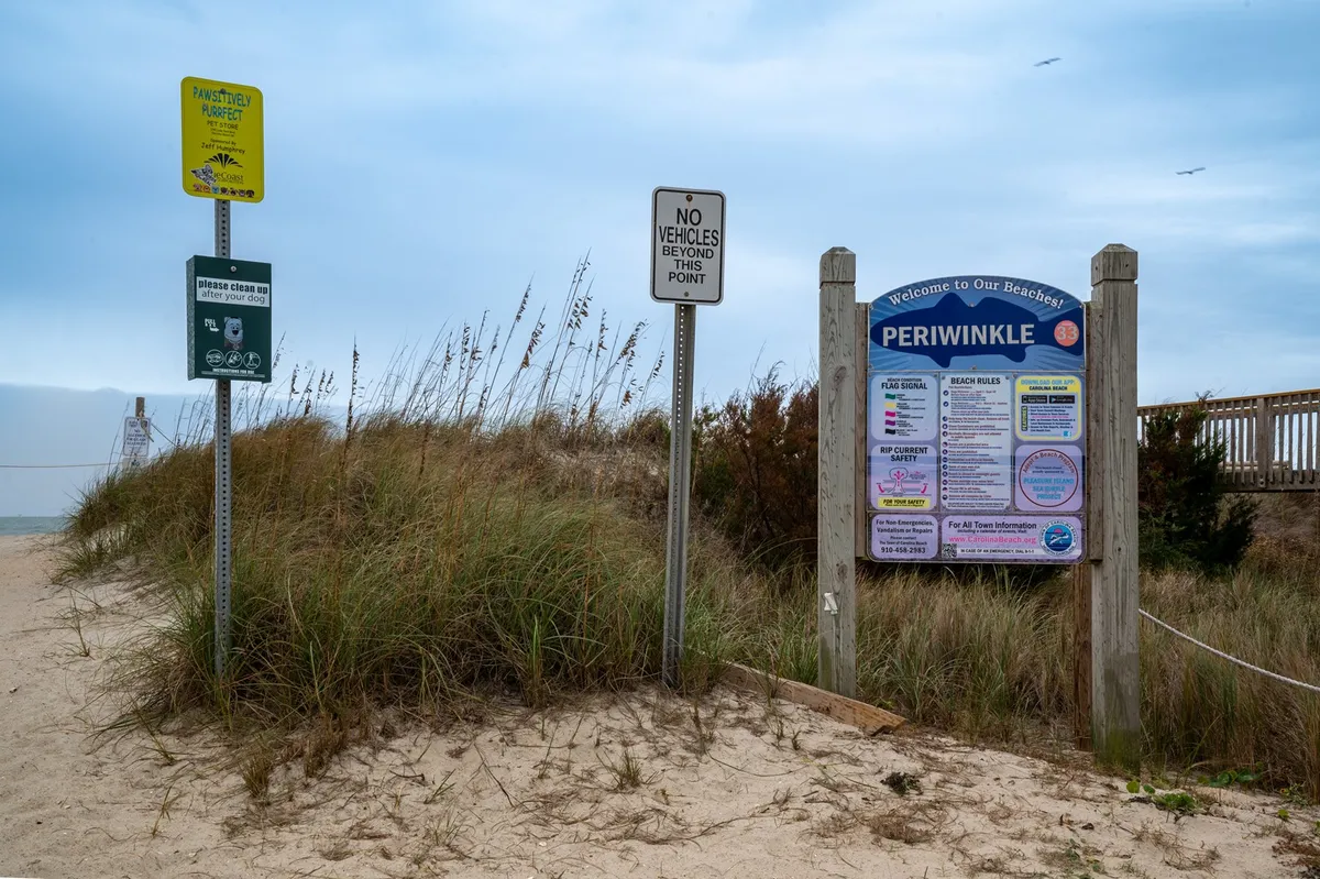 A sunny exterior shot featuring a beach access sign and informational signs, set against a relaxed backdrop. The image hints at easy, direct access to the beach for families, with clear indicators for activities like beach walking, picnicking, and swimming.