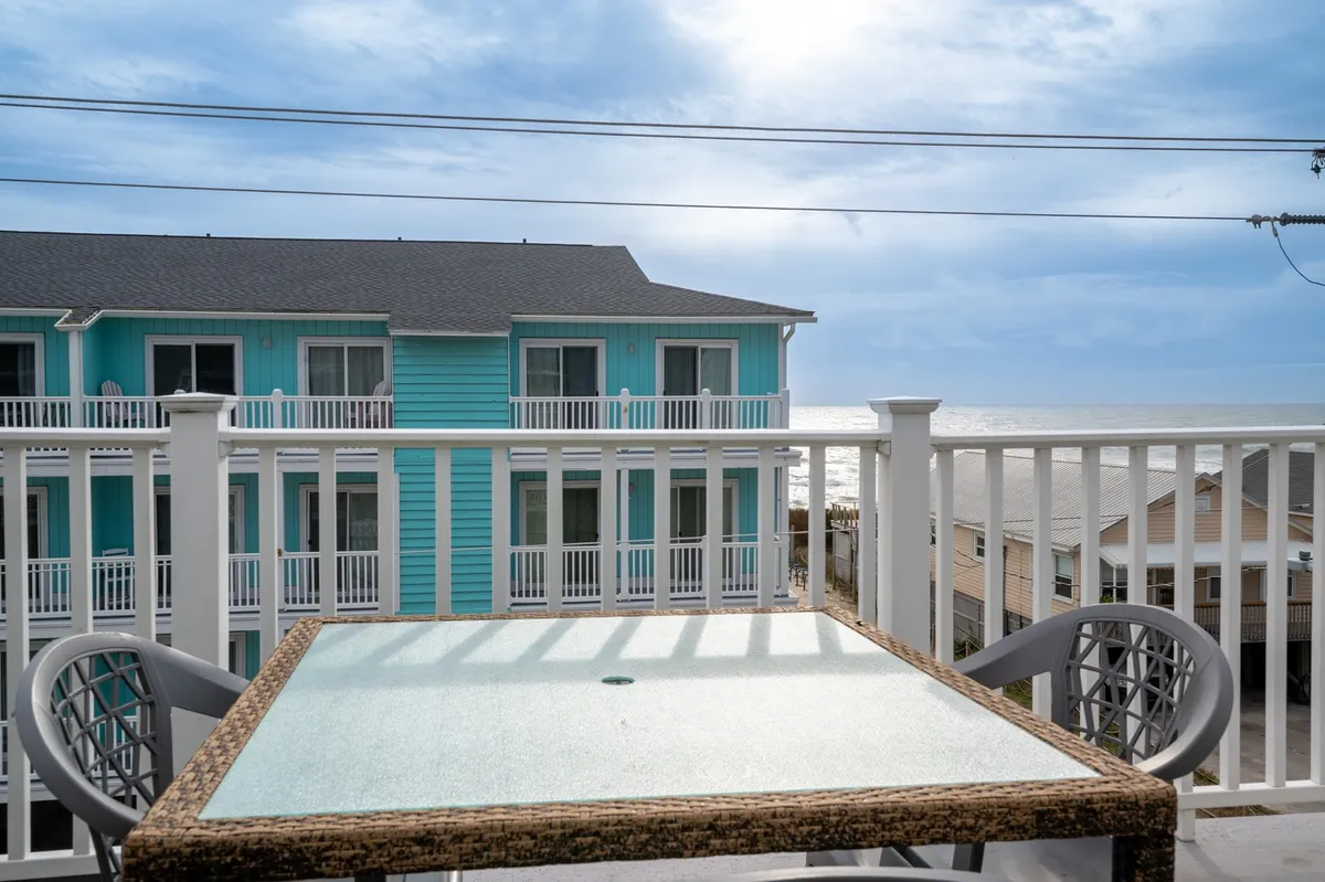 A balcony with a glass table overlooking the blue building and ocean beyond, suggesting a refreshing space for relaxing, dining, or simply enjoying views of the Atlantic.