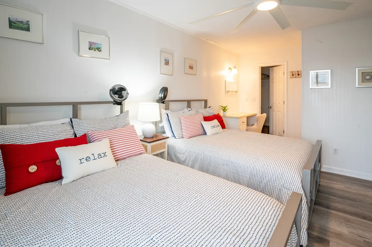 This image features two double beds adorned with decorative pillows, a nightstand with a lamp for ambient lighting, a fan on the wall, and tastefully framed artwork. The setting is calm and inviting, with plenty of space for relaxation or socializing within the primary suite.