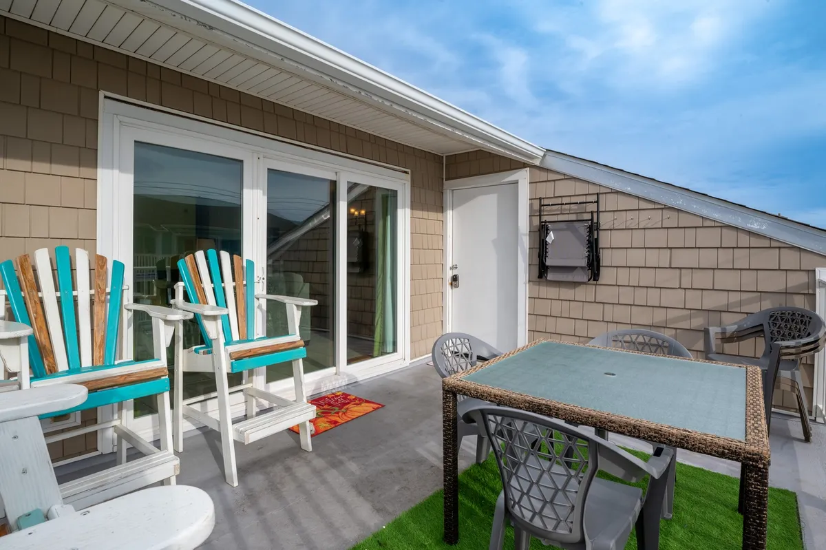 A colorful and casual balcony setting with vibrant chairs and a glass table placed on a green rug. The space is designed for outdoor dining or socializing, with an inviting atmosphere and hints of ocean or open-air views in the background. The playful aesthetic encourages gathering and relaxation.