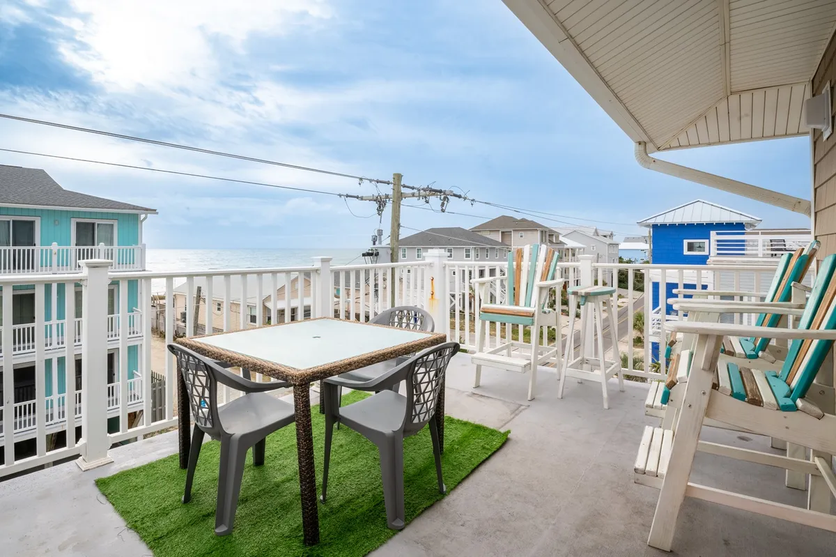 A spacious balcony featuring a table and chairs set against a backdrop of stunning ocean views, framed by a decorative railing. The outdoor furniture invites guests to dine or relax while taking in the scenery. The lighting and composition suggest the space is elevated, offering expansive vistas ideal for sunrise or sunset appreciation.