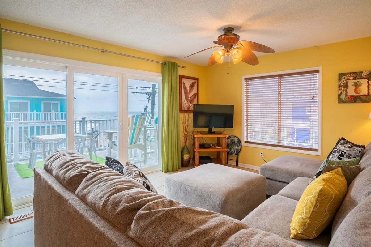 The image presents a cheerful, open living area with a large sectional sofa accented by colorful pillows. A big window offers a clear view of the beach, flooding the room with natural light. A television is positioned for easy viewing from the seating area, and decorative artwork adds a personal, welcoming touch.