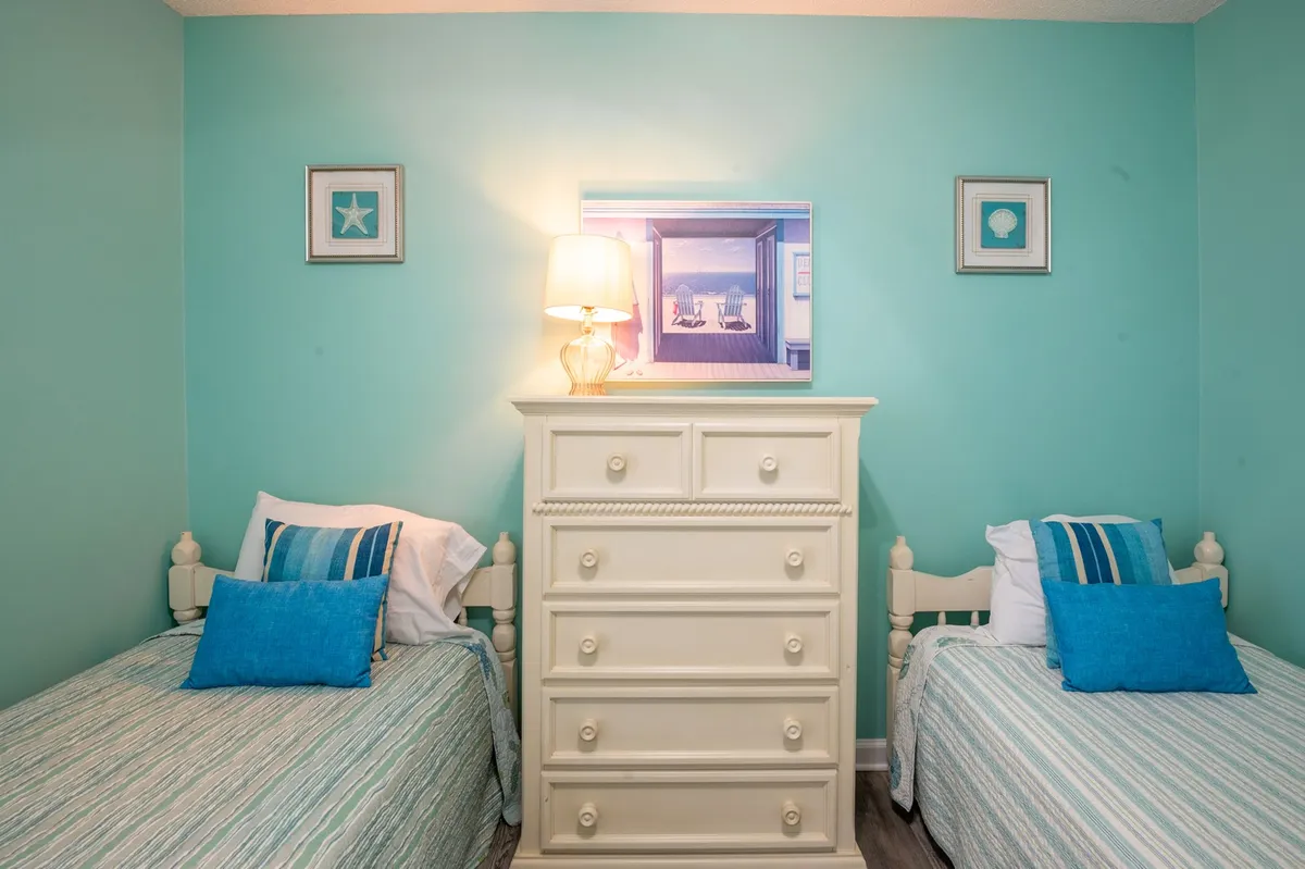 A guest bedroom with light blue walls, two twin beds dressed in soft bedding and accented with blue pillows, a white dresser, and framed artwork on the wall. The gentle morning light filters in, creating a cozy and serene retreat—perfect for children or friends.