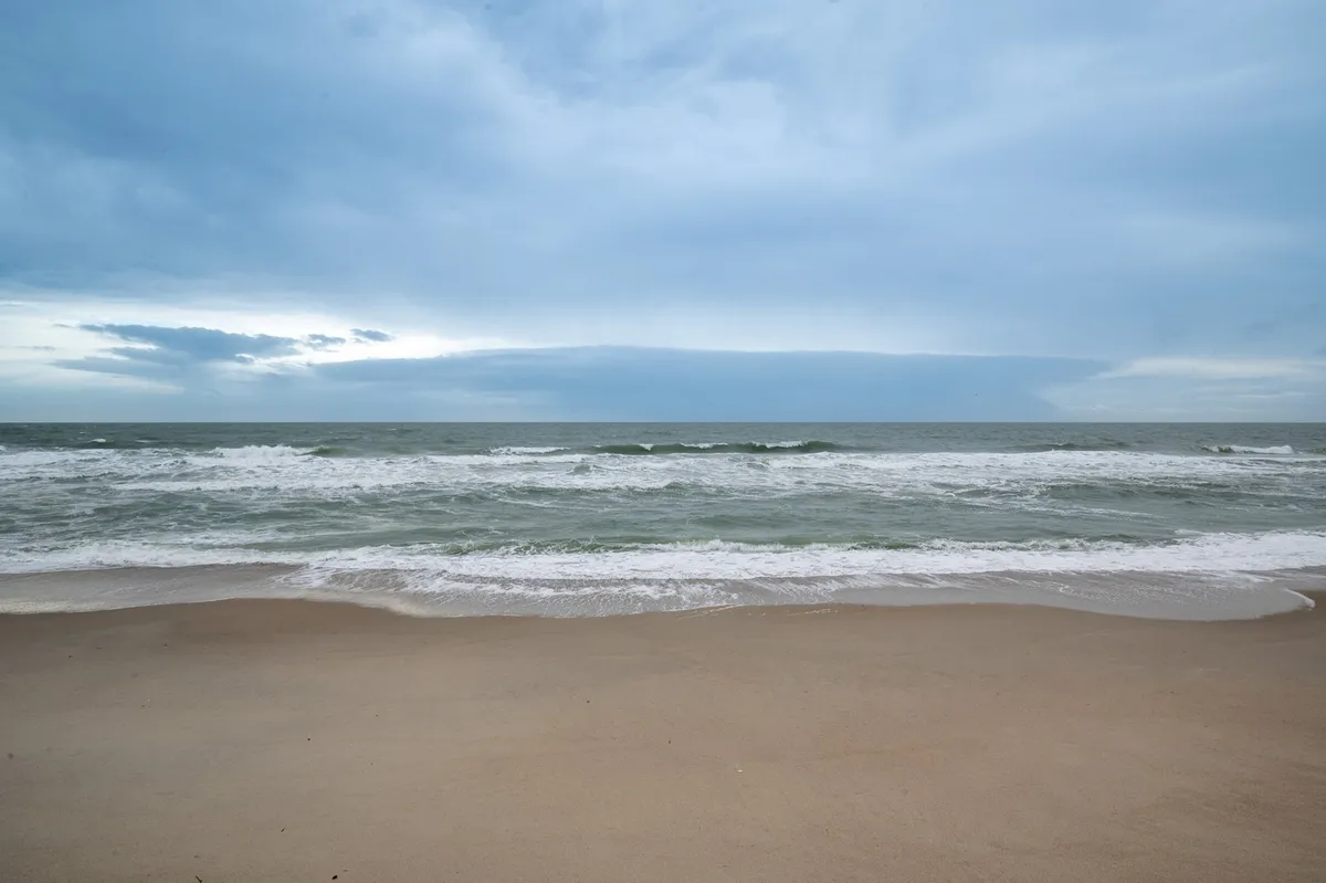 This exterior image captures the expansive sandy beach with gentle ocean waves rolling in under an overcast sky. The scene is serene, with the beach appearing inviting for calm walks or quiet contemplation, perfectly evoking the tranquil seaside experience mentioned in the section.