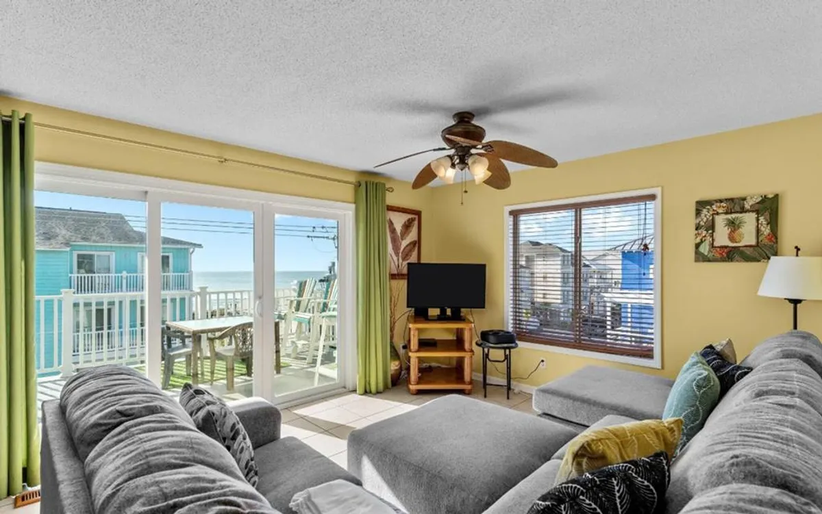The image showcases a bright, inviting living room with a plush sectional couch facing large windows that offer lovely ocean views. The space is decorated with modern wall art and includes a TV stand with a large television, making it perfect for relaxation or socializing. Natural light fills the room, creating a cozy and welcoming atmosphere.